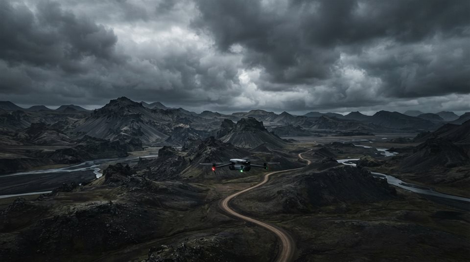 Photorealistic photo of a drone flying over a landscape, wide shot with open space at the top for text, moody dark tones, dramatic lighting, 16:9 aspect ratio