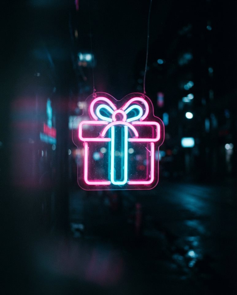 Cinematic film still of Neon glowing gift box icon with bright pink and cyan lights against a dark background, close-up detail with shallow depth of field, vivid colors, high contrast, energetic, 4:5 aspect ratio
