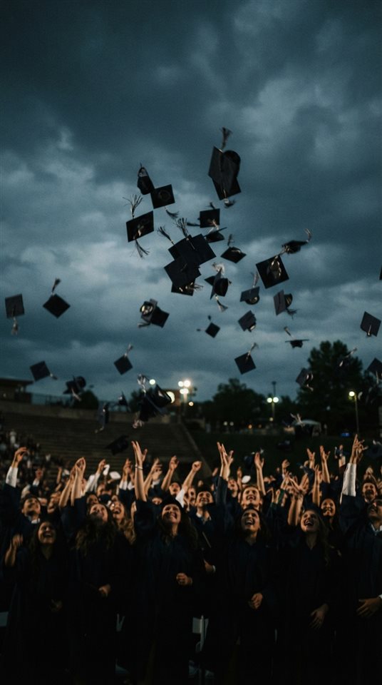 Cinematic film still of graduation caps tossed into the sky, centered subject with a simple uncluttered composition, moody dark tones, dramatic lighting, 9:16 aspect ratio