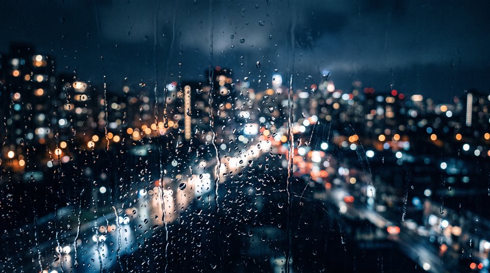 Foto fotorrealista de una ventana lluviosa por la noche con luces de la ciudad borrosas al aire libre, detalle en primer plano con poca profundidad de campo, tonos azules fríos, atmósfera silenciosa, relación de aspecto 16:9