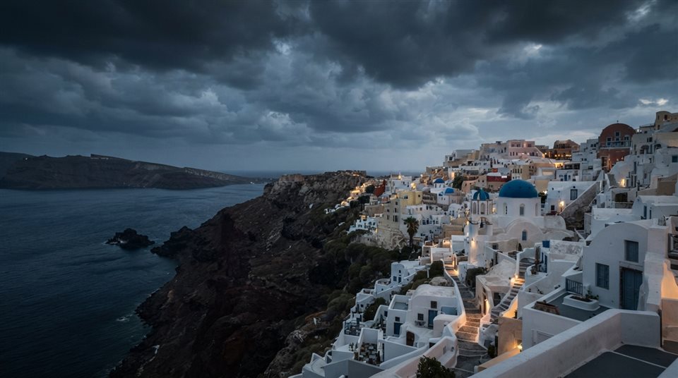 Photorealistic photo of white houses of Santorini above the sea, wide shot with open space at the top for text, moody dark tones, dramatic lighting, 16:9 aspect ratio