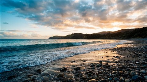 Cinematic film still of a calm wave touching a quiet pebble shore, wide shot with open space at the top for text, vivid colors, high contrast, energetic, 16:9 aspect ratio