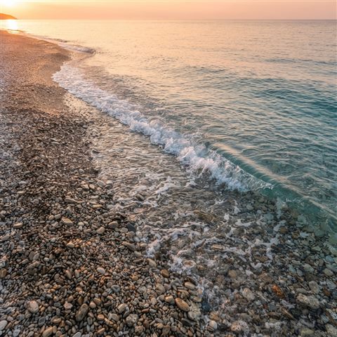 Photorealistic photo of a calm wave touching a quiet pebble shore, high-angle view from above, warm golden light, calm mood, 1:1 aspect ratio