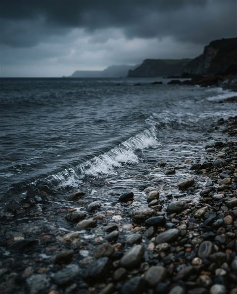 Photorealistic photo of a calm wave touching a quiet pebble shore, centered subject with a simple uncluttered composition, moody dark tones, dramatic lighting, 4:5 aspect ratio