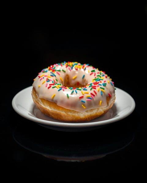 Photorealistic photo of A sweet glazed ring donut topped with colorful sprinkles, resting on a clean white plate., wide shot with open space at the top for text, moody dark tones, dramatic lighting, 4:5 aspect ratio