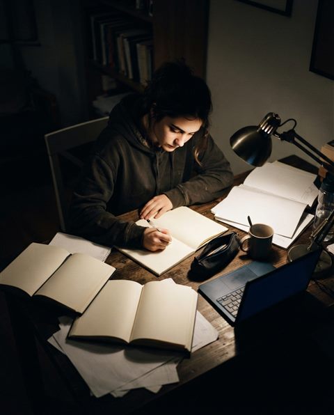 Photorealistic photo of a student studying late at night under a desk lamp, high-angle view from above, moody dark tones, dramatic lighting, 4:5 aspect ratio