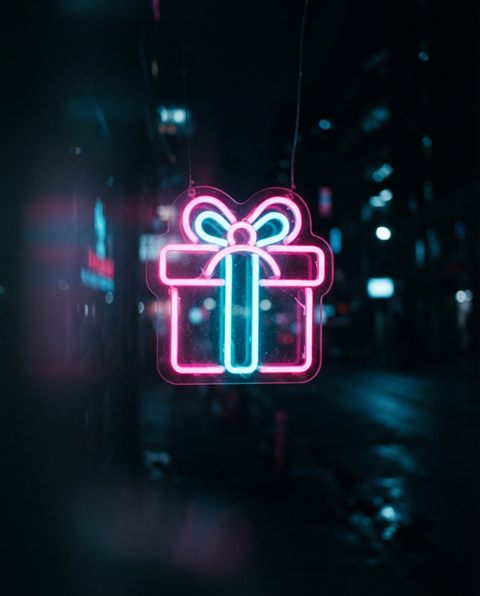 Cinematic film still of Neon glowing gift box icon with bright pink and cyan lights against a dark background, close-up detail with shallow depth of field, vivid colors, high contrast, energetic, 4:5 aspect ratio