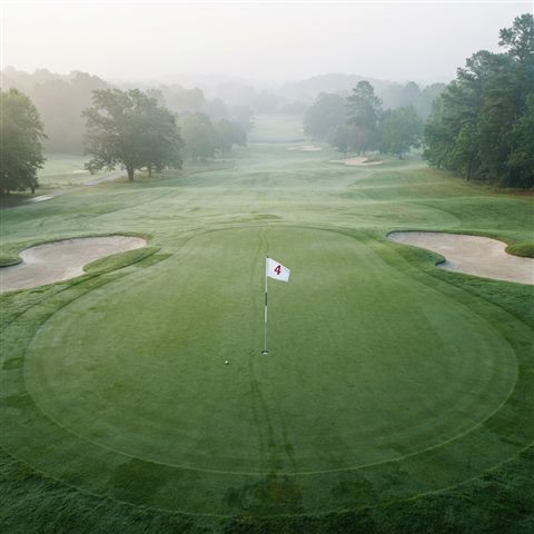 Photorealistic photo of a golf course green with a flag, high-angle view from above, misty morning haze, dreamy, 1:1 aspect ratio