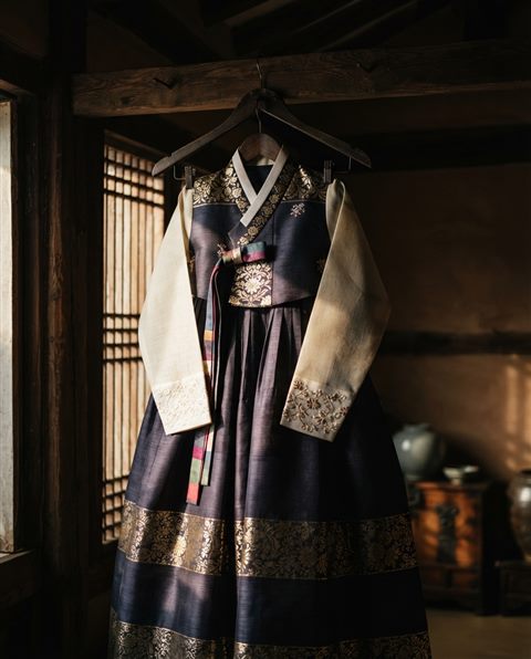 Photorealistic photo of a traditional Korean hanbok dress displayed on a wooden hanger in a sunlit hanok room, close-up detail with shallow depth of field, moody dark tones, dramatic lighting, 4:5 aspect ratio