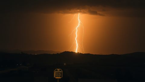 Photorealistic photo of A jagged bolt of lightning flashing across a dark night sky over a silhouette of hills, high-angle view from above, warm golden light, calm mood, 16:9 aspect ratio