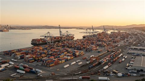 Photorealistic photo of a container shipping port viewed from afar with stacked cargo boxes, wide shot with open space at the top for text, warm golden light, calm mood, 16:9 aspect ratio