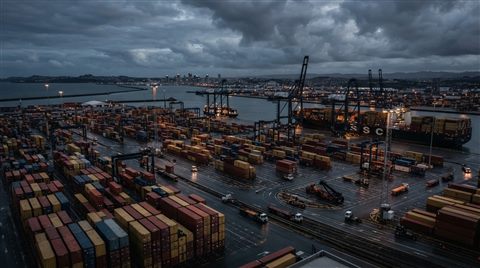 Photorealistic photo of a container shipping port viewed from afar with stacked cargo boxes, high-angle view from above, moody dark tones, dramatic lighting, 16:9 aspect ratio