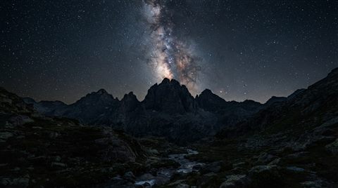 Photorealistic photo of milky way over mountains, centered subject with a simple uncluttered composition, moody dark tones, dramatic lighting, 16:9 aspect ratio