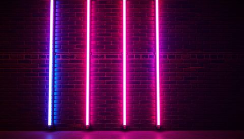 Photorealistic photo of Vibrant pink and blue glowing neon light tubes creating an abstract urban night atmosphere on a dark brick wall., centered subject with a simple uncluttered composition, warm golden light, calm mood, 16:9 aspect ratio
