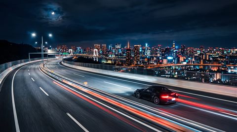Photorealistic photo of car driving on an empty highway at night with city lights, wide shot with open space at the top for text, vivid colors, high contrast, energetic, 16:9 aspect ratio