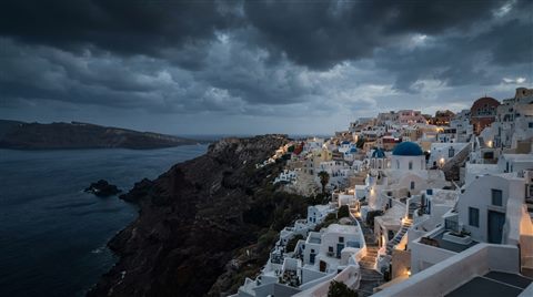 Photorealistic photo of white houses of Santorini above the sea, wide shot with open space at the top for text, moody dark tones, dramatic lighting, 16:9 aspect ratio