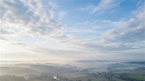 Photorealistic photo of blue sky with soft clouds, wide shot with open space at the top for text, misty morning haze, dreamy, 16:9 aspect ratio
