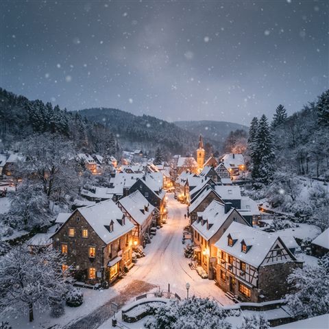 Photorealistic photo of a cozy small village covered in fresh white snow during a gentle snowfall, wide shot with open space at the top for text, warm golden light, calm mood, 1:1 aspect ratio
