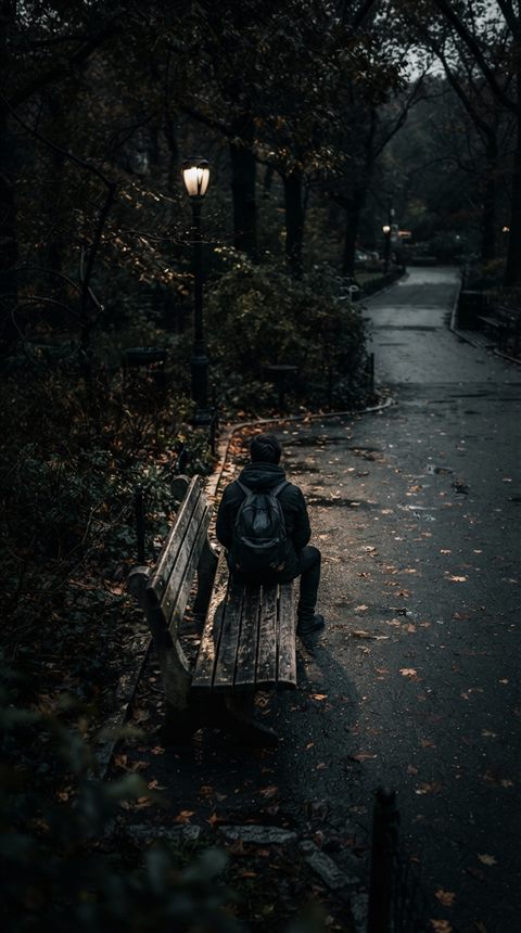 Photorealistic photo of person sitting alone on a park bench seen from behind, high-angle view from above, moody dark tones, dramatic lighting, 9:16 aspect ratio