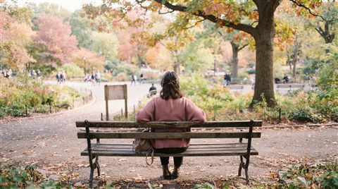 Cinematic film still of person sitting alone on a park bench seen from behind, high-angle view from above, soft pastel colors, bright and cozy, 16:9 aspect ratio