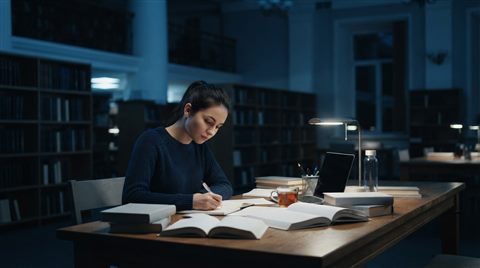 Photorealistic photo of a student studying at a desk with books, wide shot with open space at the top for text, cool blue tones, quiet atmosphere, 16:9 aspect ratio