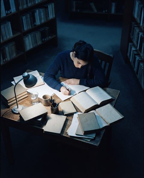 Cinematic film still of a student studying at a desk with books, high-angle view from above, cool blue tones, quiet atmosphere, 4:5 aspect ratio