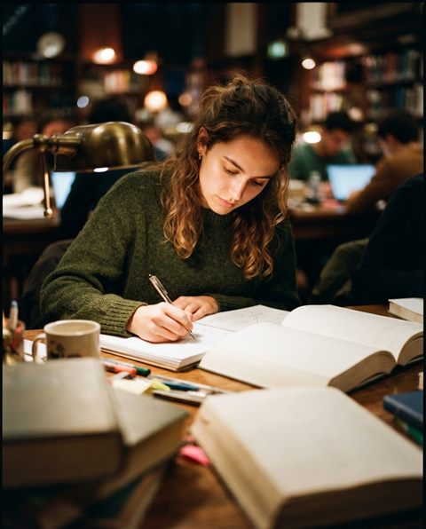 Cinematic film still of a student studying at a desk with books, close-up detail with shallow depth of field, vivid colors, high contrast, energetic, 4:5 aspect ratio